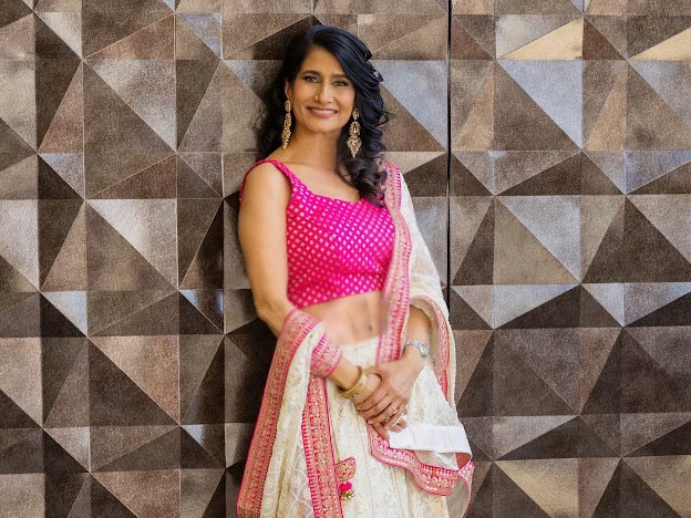 Smiling woman in pink traditional dress with a shawl, standing against a geometric patterned wall.