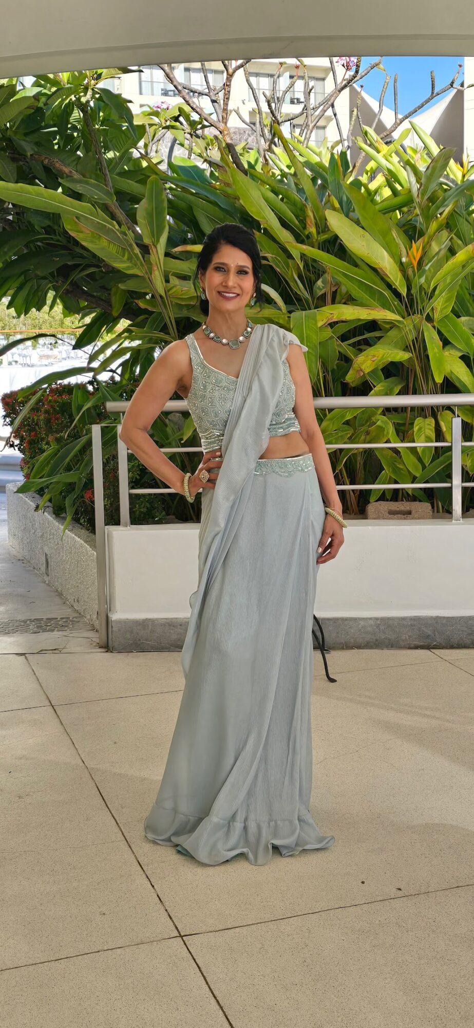 Woman in a light-colored dress standing outdoors with large green plants in the background.
