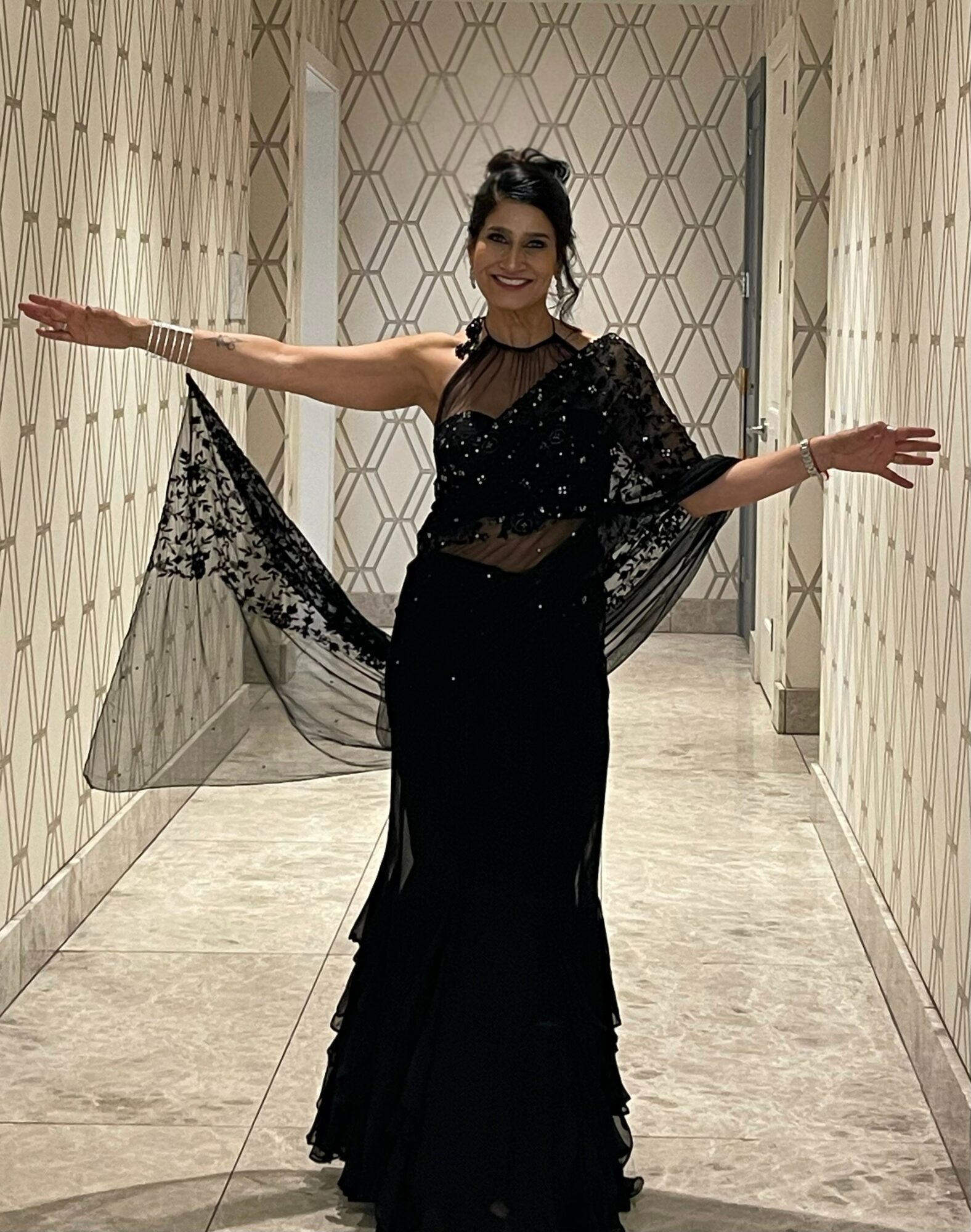 Woman in black dress with lace shawl smiling with arms outstretched in a hallway.