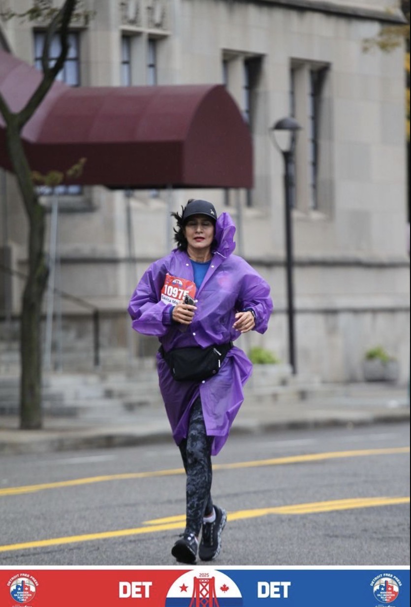 Woman walking on street holding a coffee cup, wearing a purple raincoat, black cap, and patterned leggings, with buildings in background.