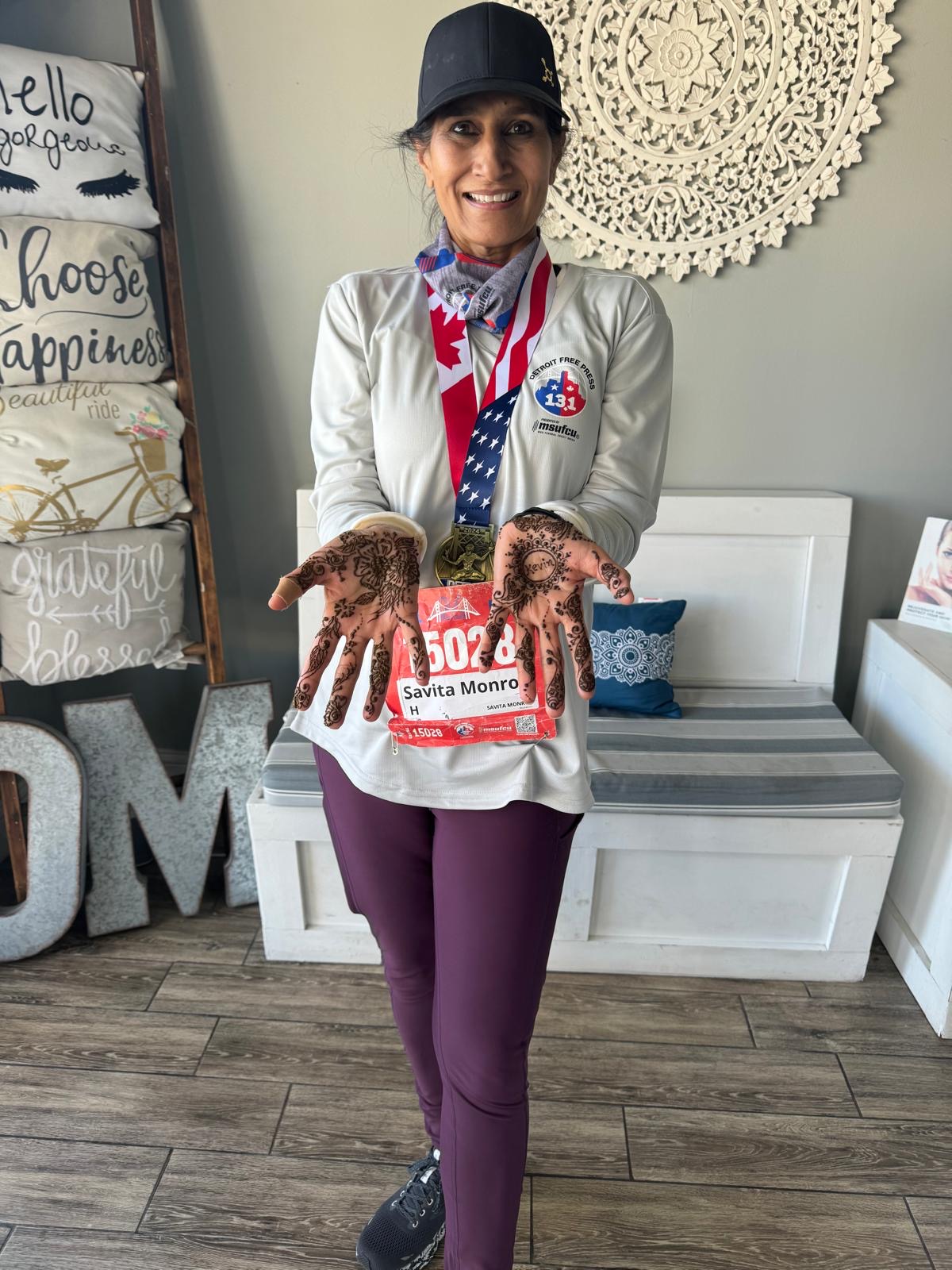 Woman smiling, wearing a black cap, gray jacket, and purple pants, holding a race bib with her hands covered in mud, standing indoors.