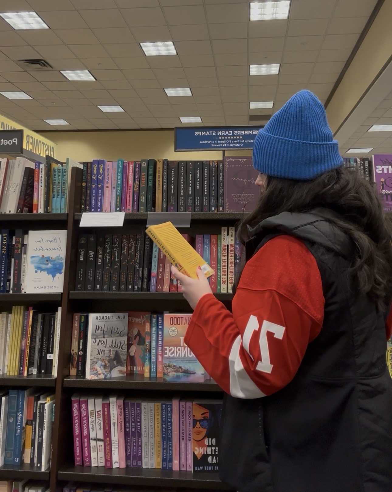 Person wearing a blue beanie and red jacket browsing books in a bookstore, holding a yellow book.