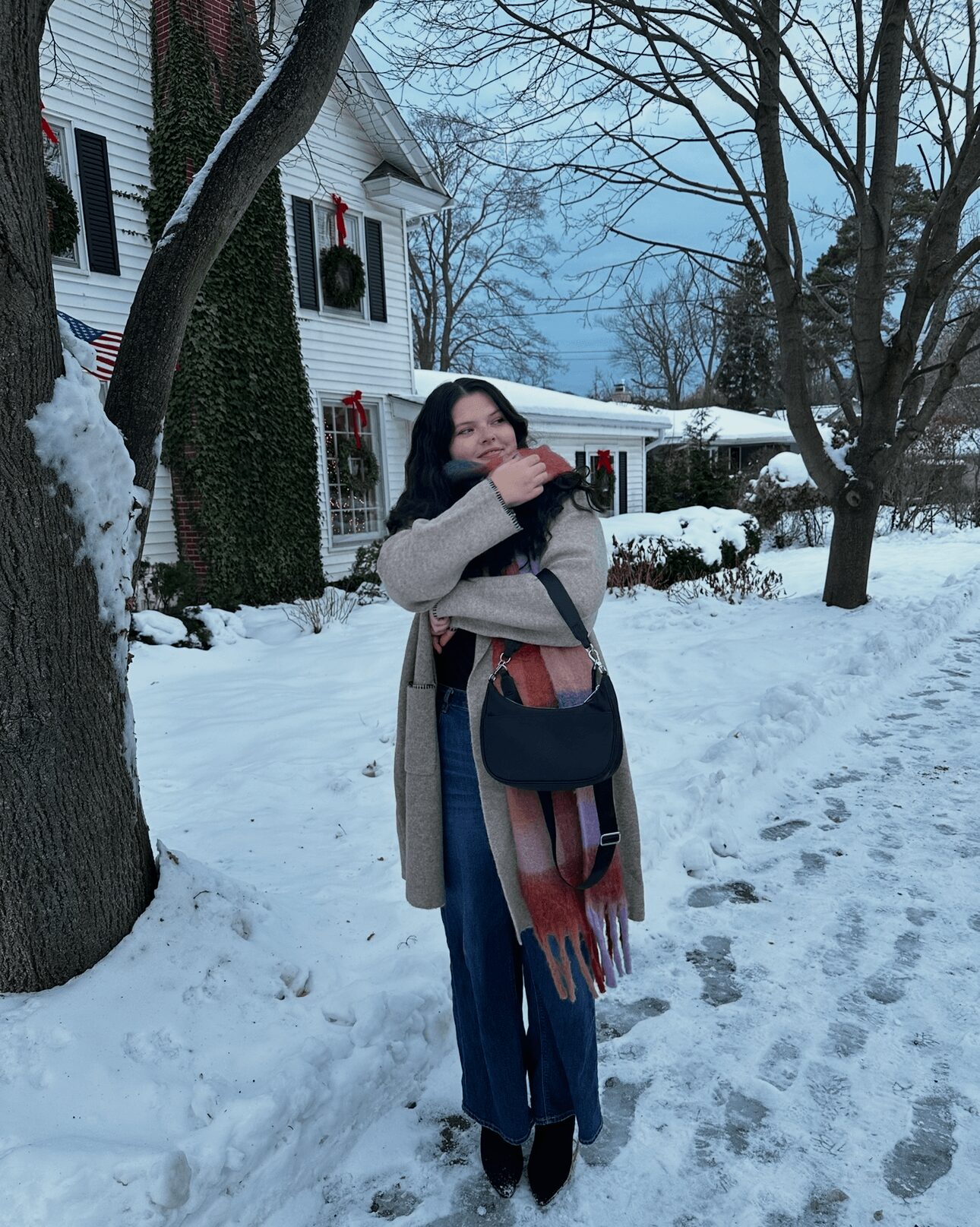 Woman standing outdoors in snow, wearing a coat and scarf, with a house decorated for winter in the background.