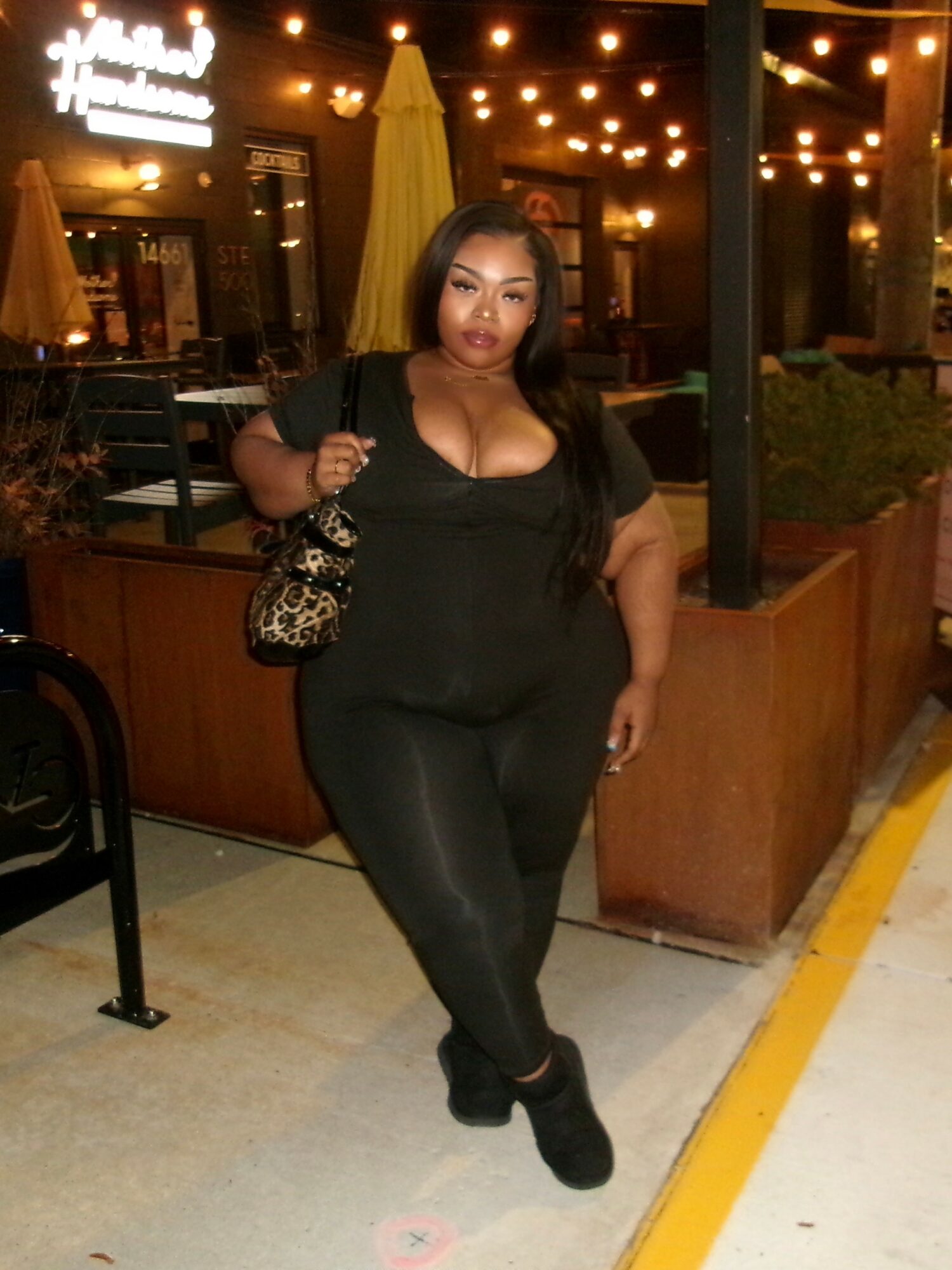 Woman in black outfit standing outside at night with string lights overhead, holding a patterned bag, in a restaurant patio area.