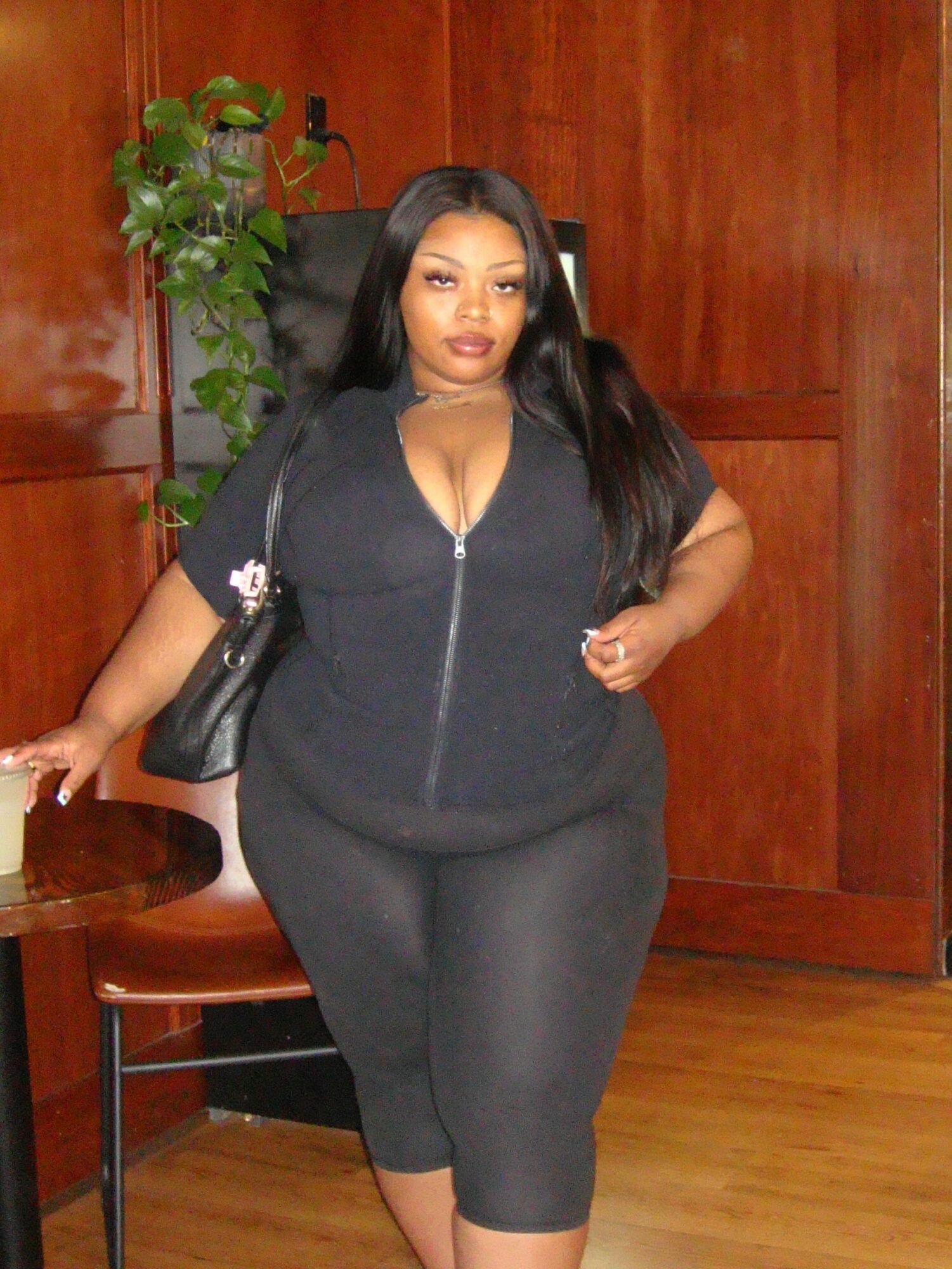 Woman with long black hair in black outfit standing in a room with wooden walls and a plant, posing with hand on hip.