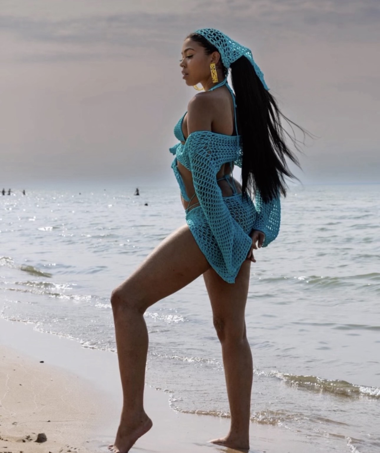 Woman standing on beach with ocean in background, wearing blue crochet cover-up and headscarf, looking to the side.