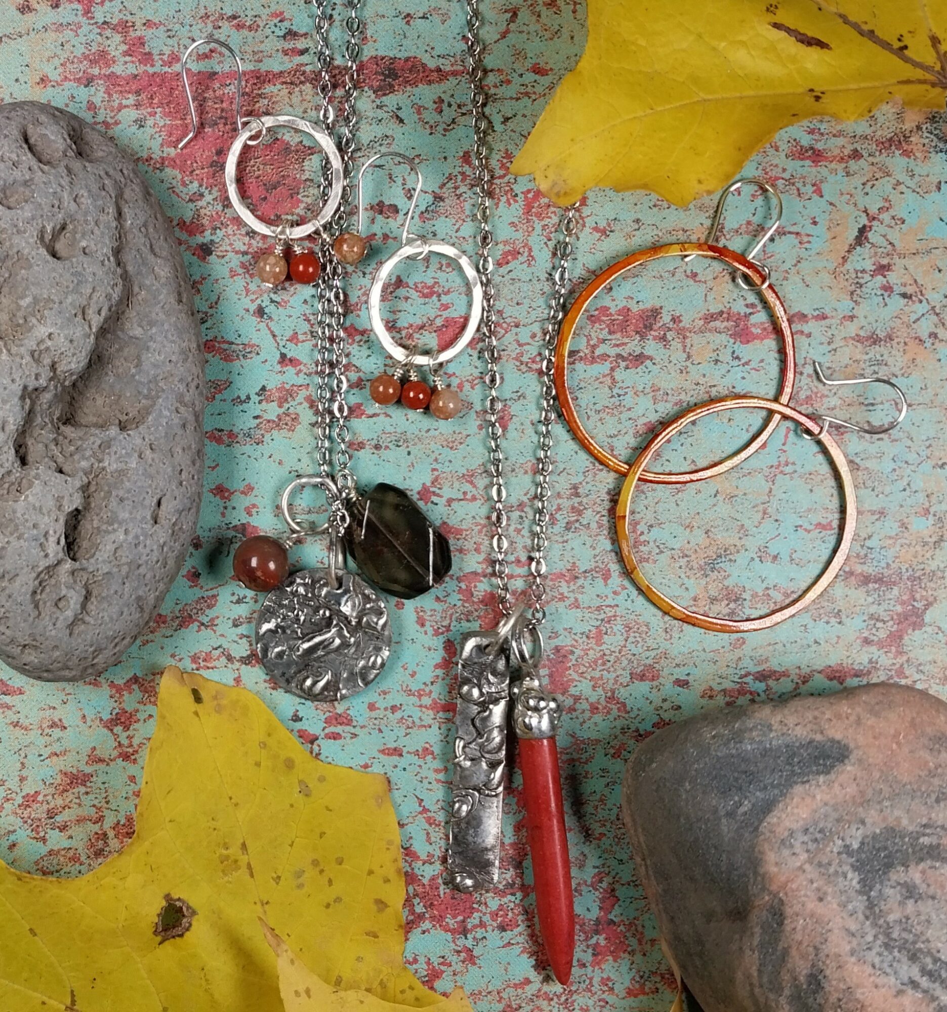 Jewelry including earrings, necklaces, and bracelets on a weathered wooden surface with yellow leaves and rocks.