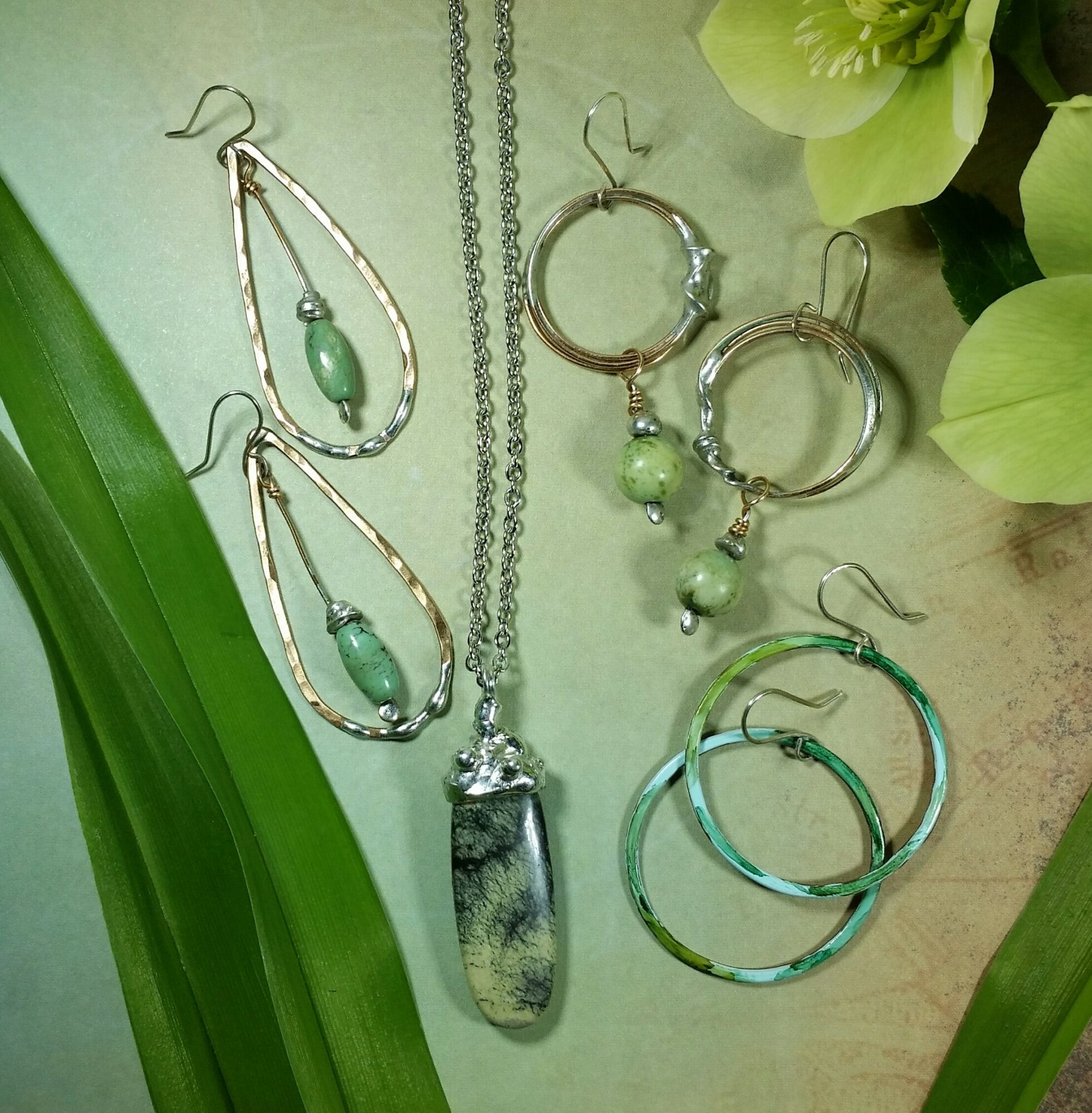 Jewelry including earrings, necklace, and rings with green stones and silver elements, arranged on a light background with green leaves.