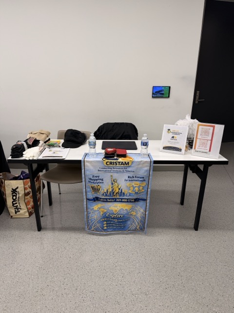 Table with bottles, papers, and a poster, against a plain wall with a small screen, in a room with gray carpet.