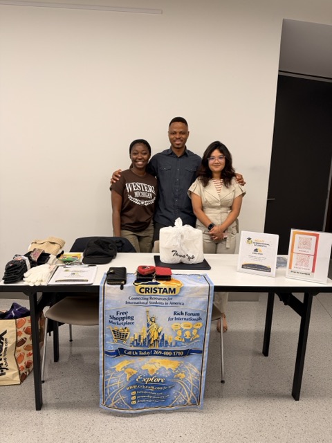 Three people standing behind a table with promotional materials, in a room with plain walls.