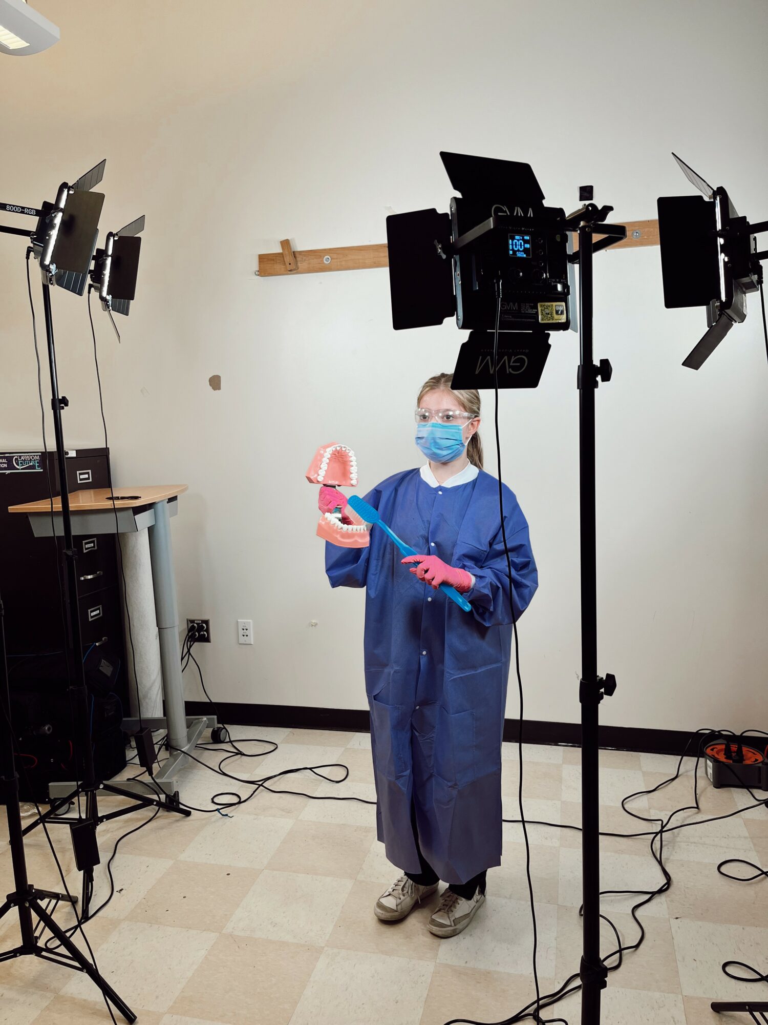 Person in blue gown and face mask holding a pink object, surrounded by studio lighting equipment, in a room with white walls.