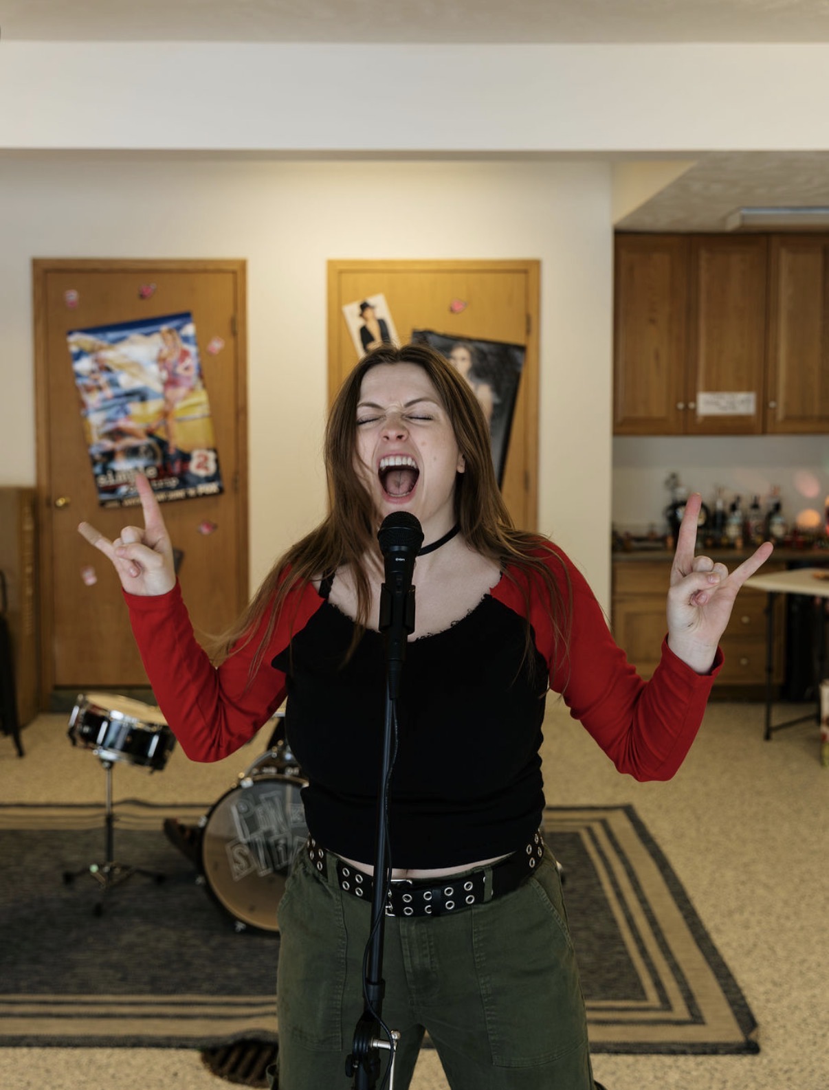 Young woman singing passionately into a microphone with eyes closed and hands raised, in a room with musical instruments and artwork.