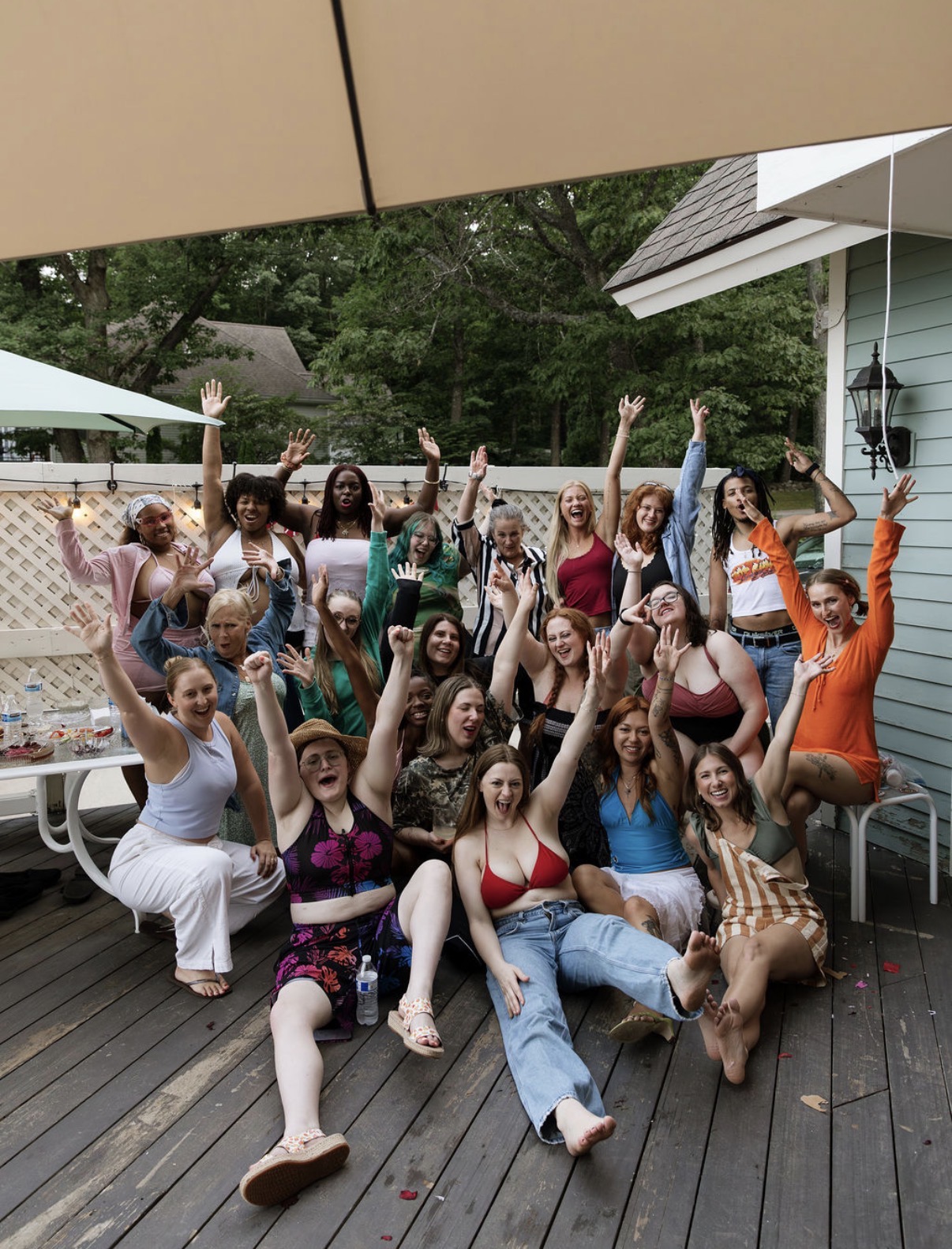 Group of people celebrating outdoors on a wooden deck, raising their hands, with trees and a house in the background.