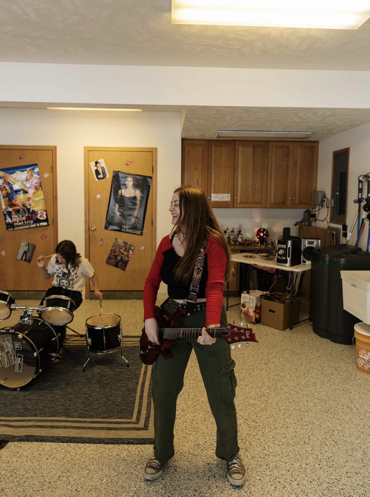 Person with long hair holding a guitar in a room with drums, posters, and wooden cabinets.