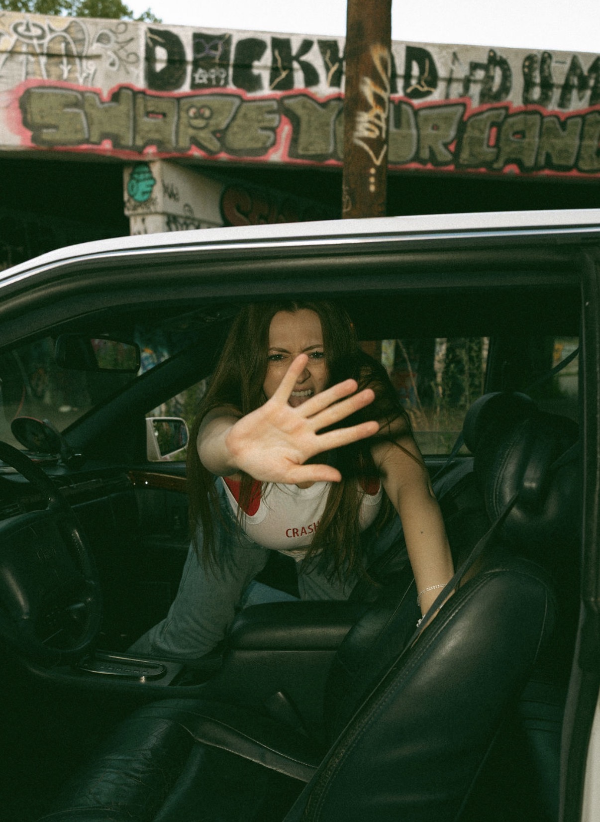 Person inside a vehicle with hand raised, graffiti-covered wall in background, partially visible face, dark hair, casual clothing.
