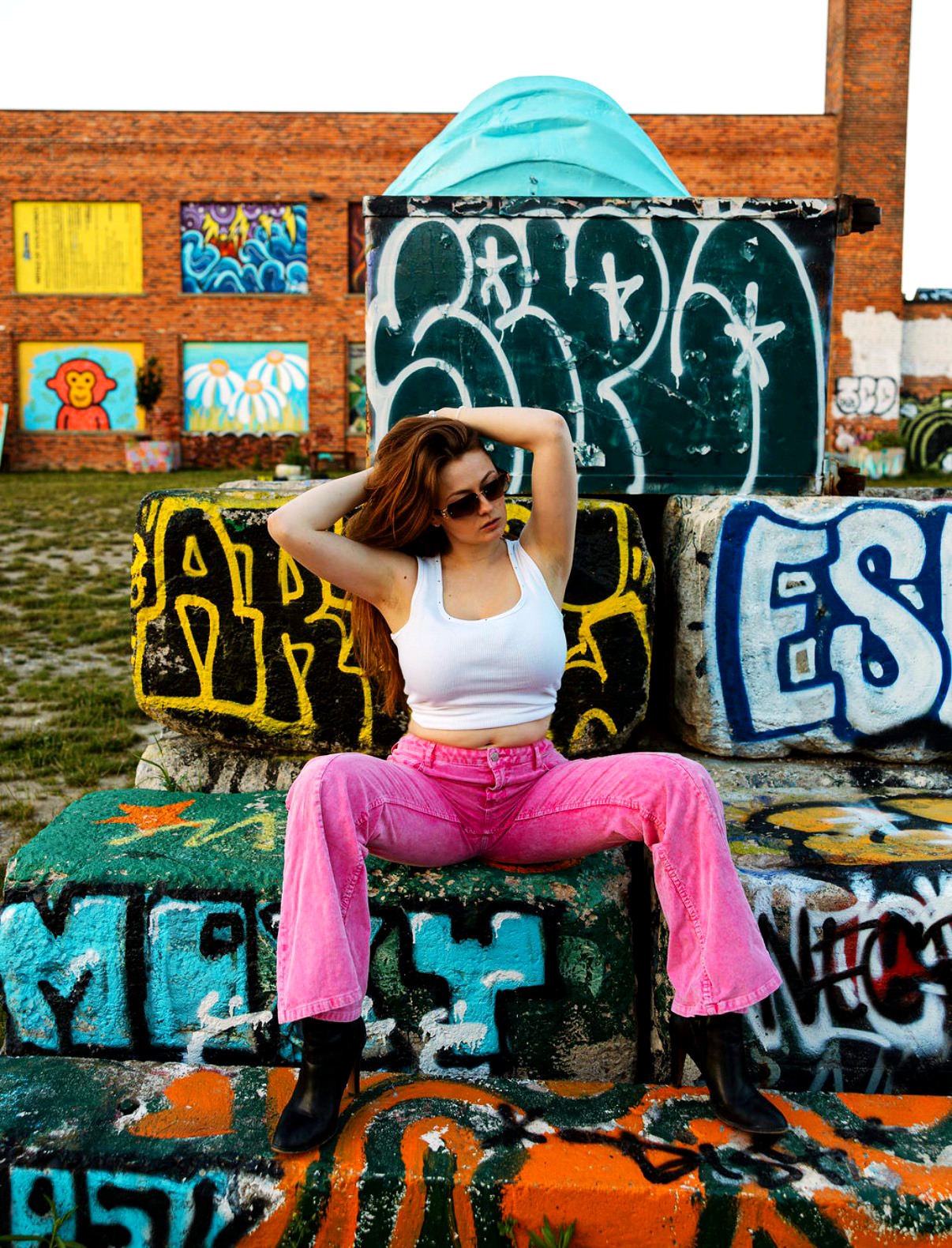 Woman with sunglasses sitting on graffiti-covered blocks, wearing a white top and pink pants, with her hands behind her head.