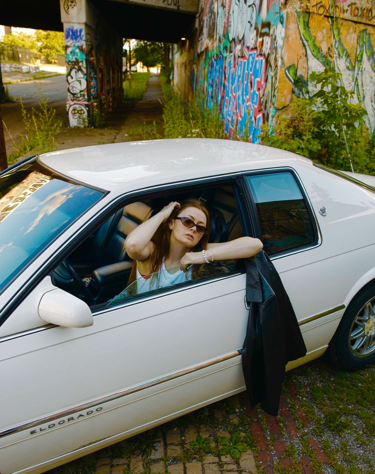 Woman with sunglasses leaning out of a white car window, arm resting on the door, graffiti-covered wall in background.