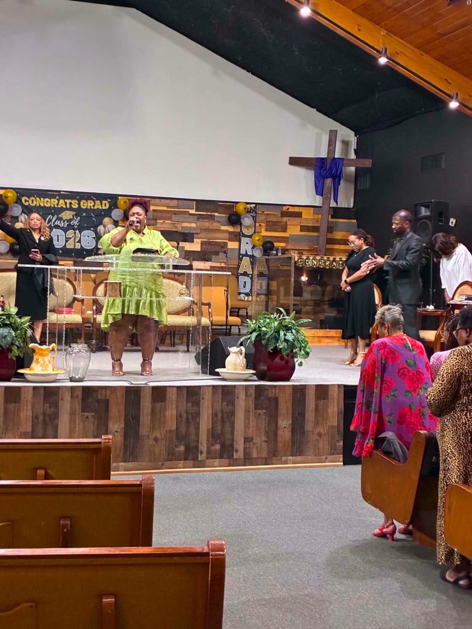 Group of people on stage, some singing or speaking, with a decorated background and audience in foreground, in a church or event hall.