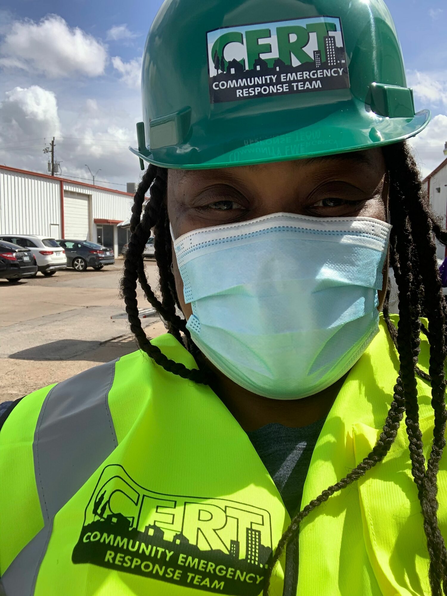 Person wearing a green helmet and high-visibility vest, with a face mask, outdoors in a parking lot.