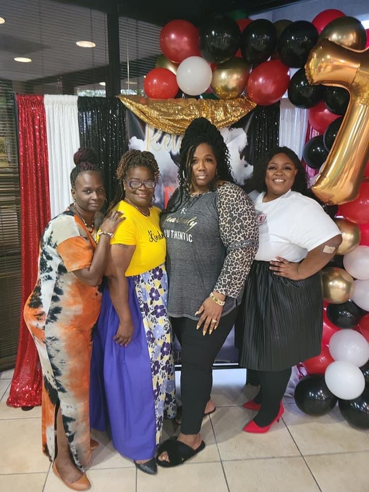 Four women standing together in front of a decorated backdrop with balloons, celebrating a special occasion, smiling at the camera.