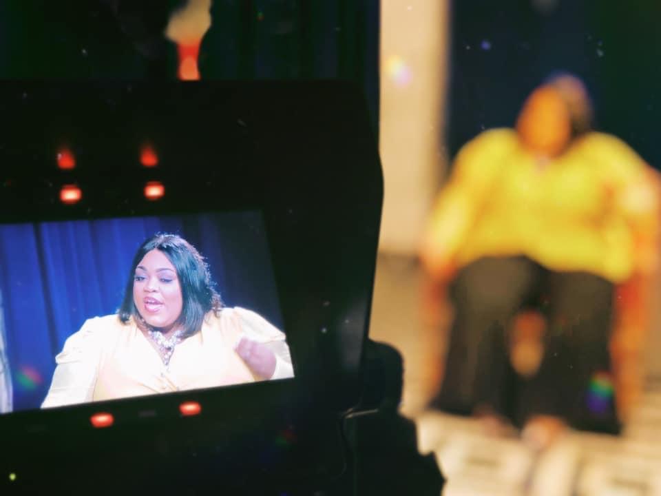 Television monitor shows a woman speaking, with a blurred person in yellow in the background.