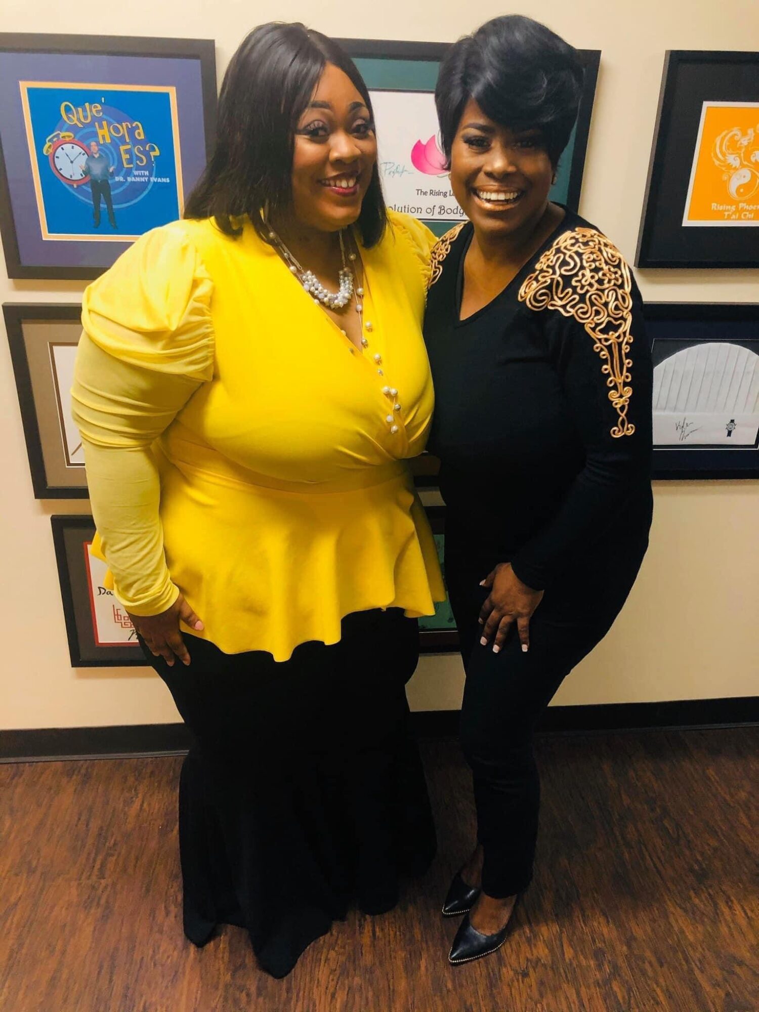 Two women standing close together, smiling, in front of colorful framed artwork on a wall.