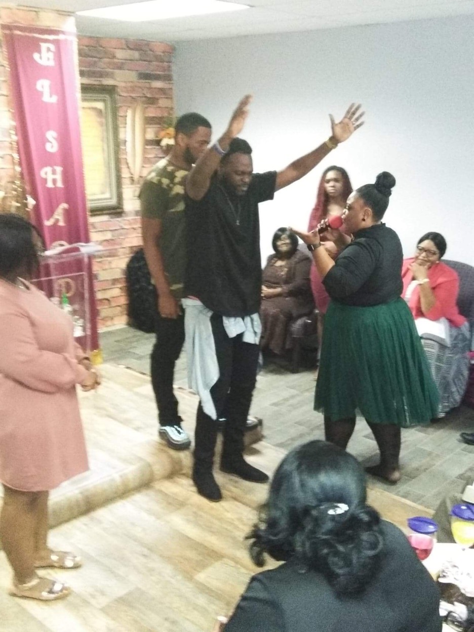 Group of people in a room, some standing and some seated, with a person raising hands in the center.