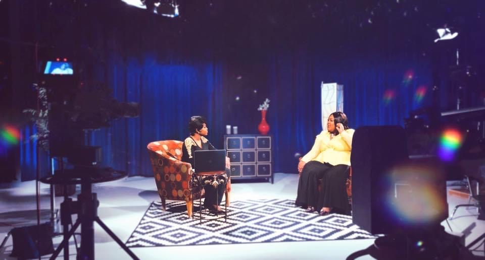 Two women sit and talk on a TV set with blue curtains, a patterned rug, and studio lights.