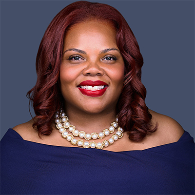Woman with red hair, wearing a navy blue top and pearl necklace, smiling against a gray background.