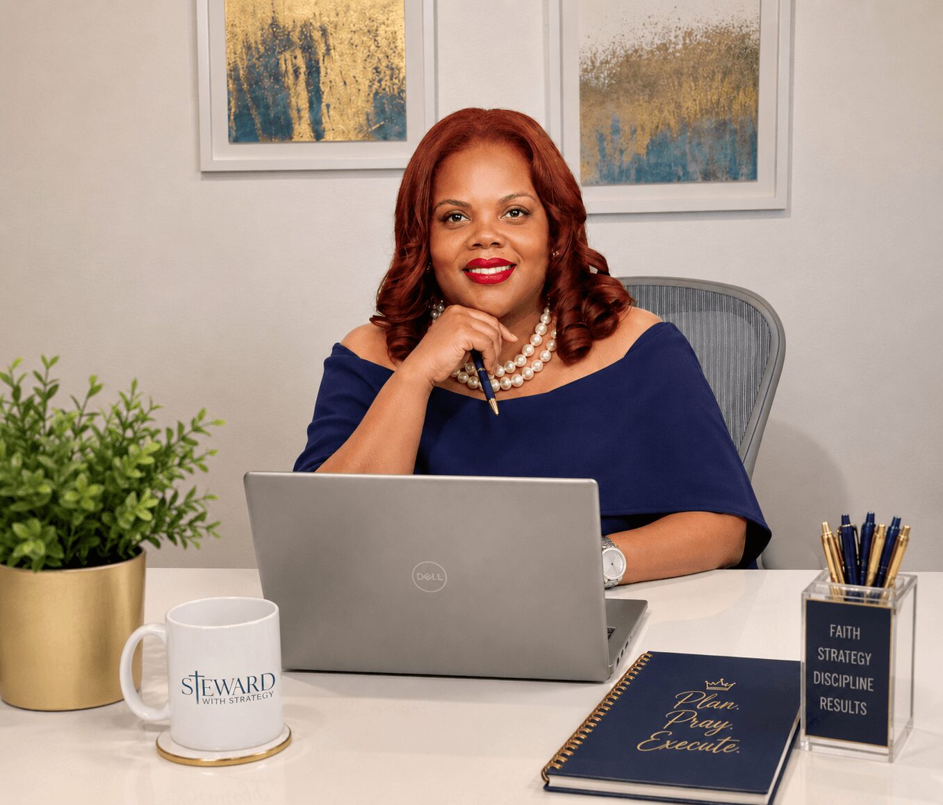Woman with red hair and pearl necklace sitting at desk with laptop, plant, mug, and notebook, smiling, in office setting.