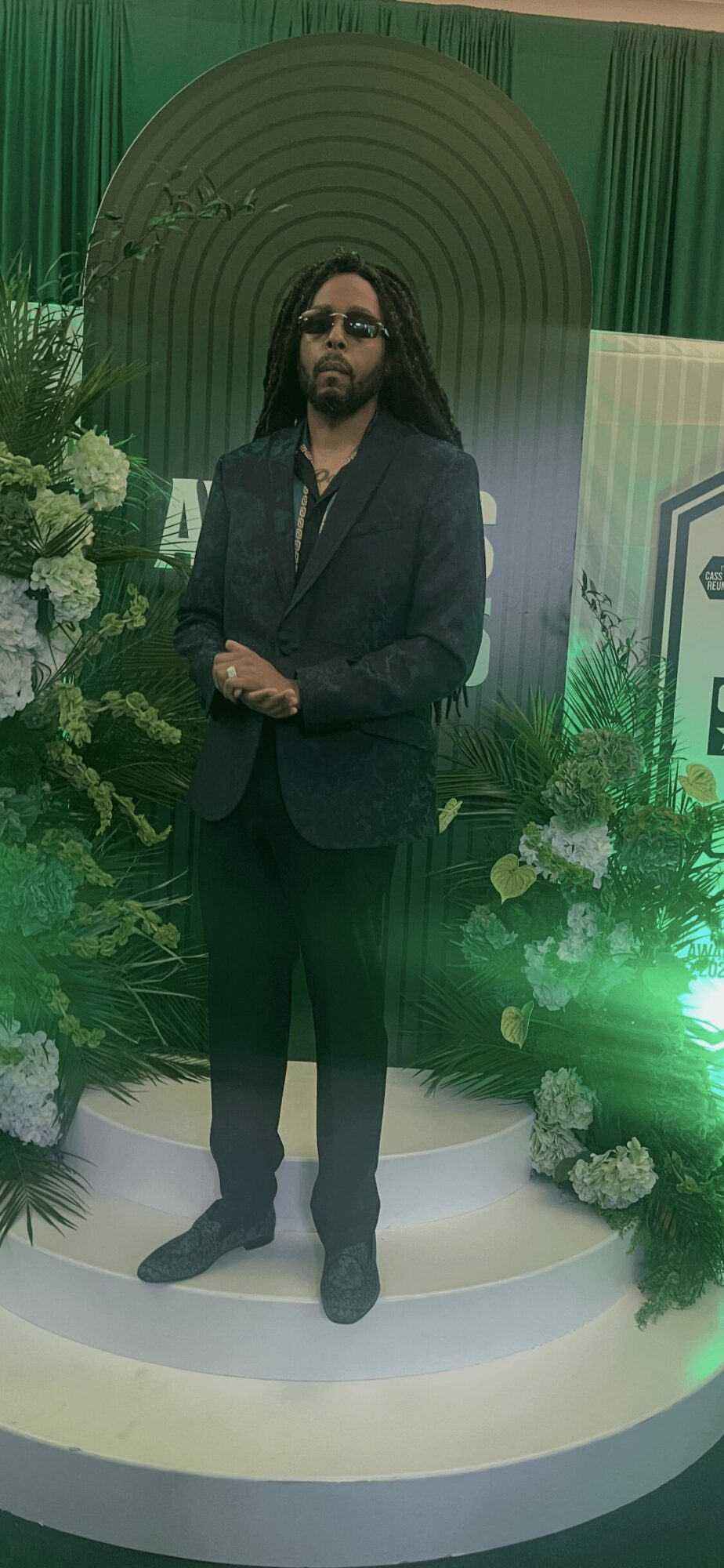 Man with long hair and sunglasses in a dark suit standing on a decorated stage with flowers and green lighting.