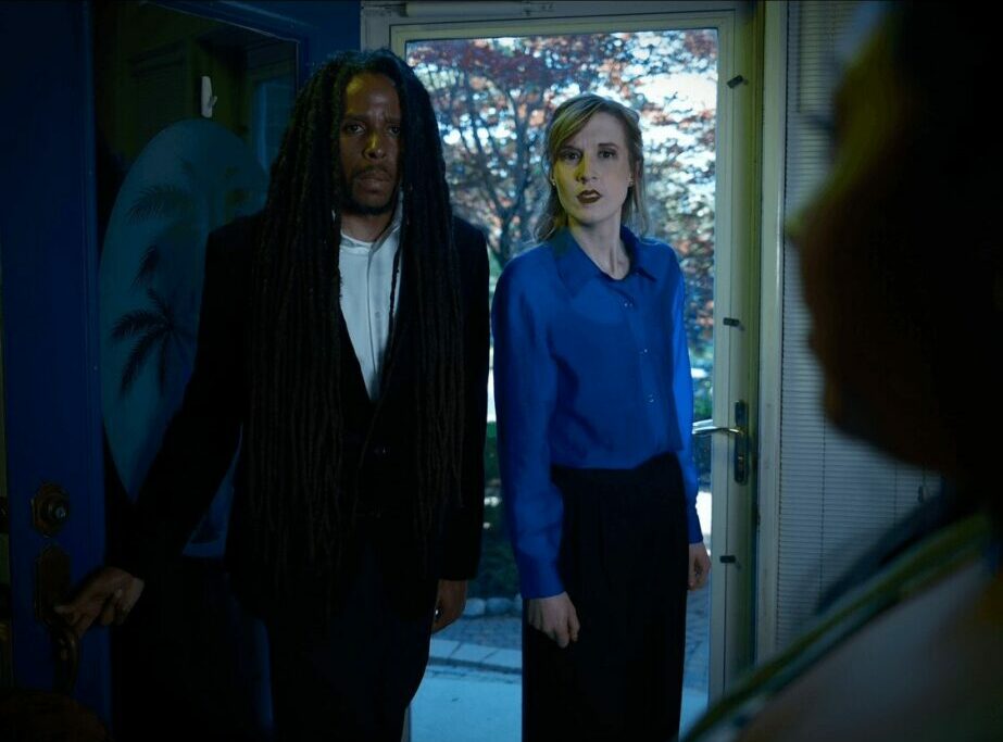Two women stand indoors near a window, with a third person partially visible in the foreground, in a dimly lit room.