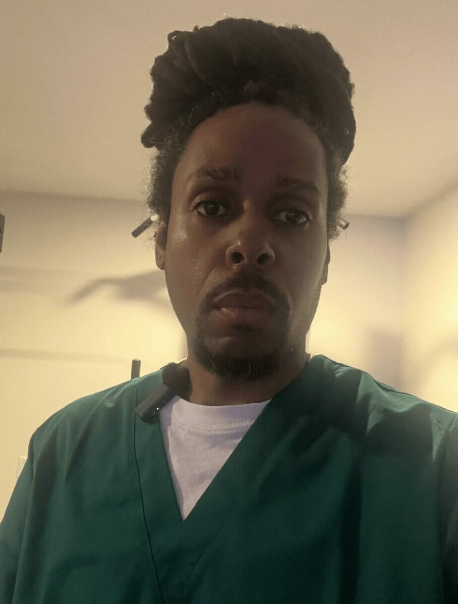 Person with dark curly hair wearing green scrubs, looking at camera indoors, ceiling fan visible in background.