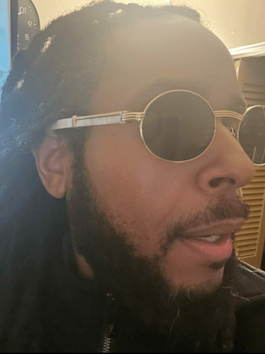 Close-up of a person wearing round sunglasses and a dark jacket, with a beard and dreadlocks, indoors.