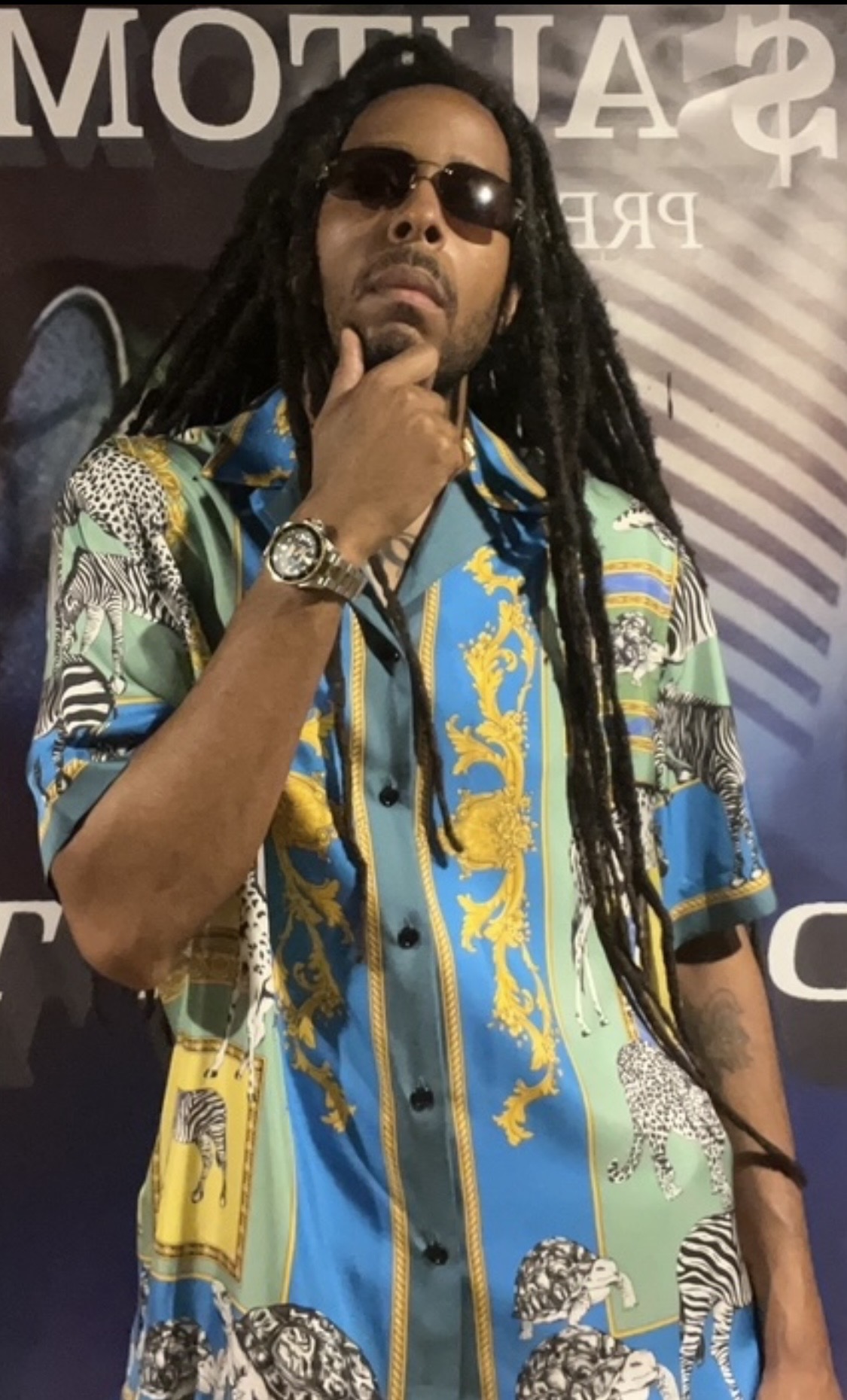 Man with long dreadlocks wearing sunglasses and a colorful patterned shirt, standing with hand on chin.