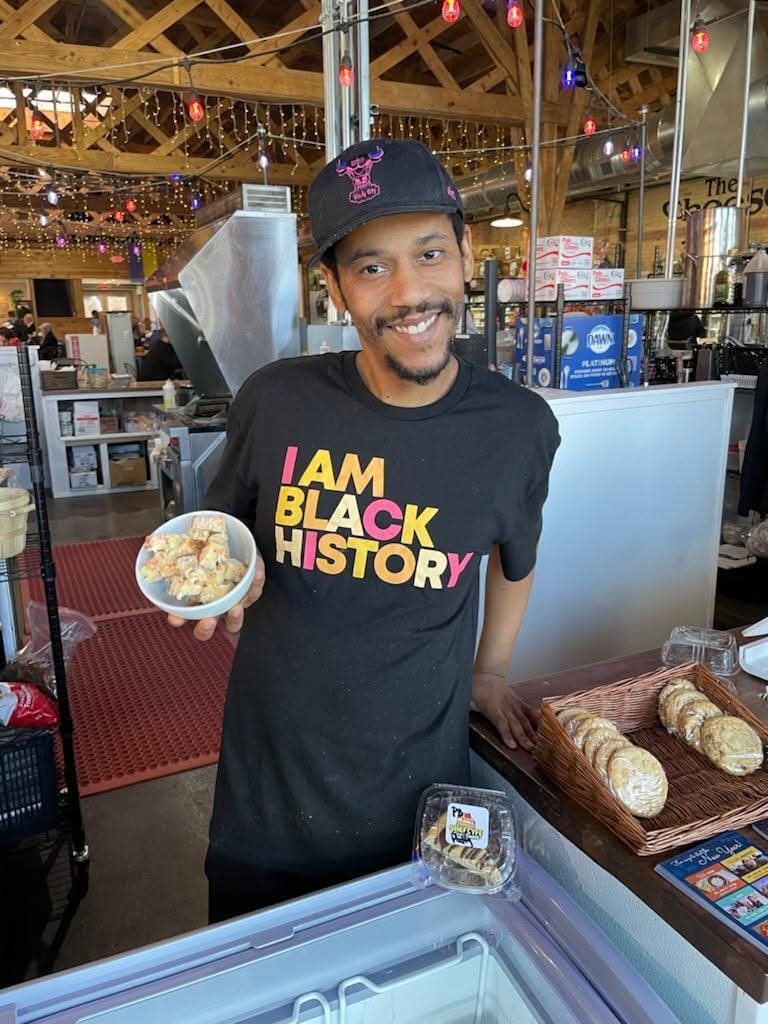 Smiling man wearing a black T-shirt with colorful text and a black cap, holding a bowl of food in a market or food hall.