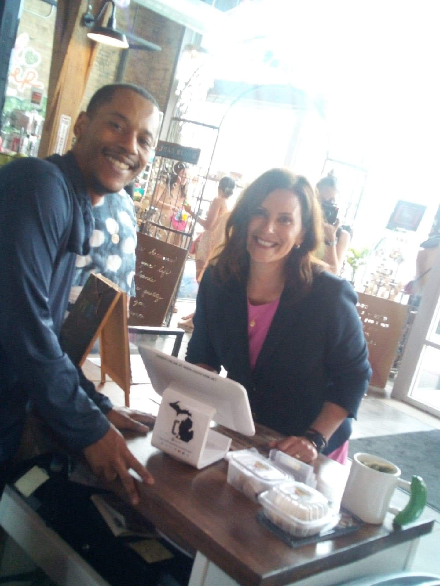 Two people smiling at a counter in a bright cafe or shop, with a window and shelves in the background.