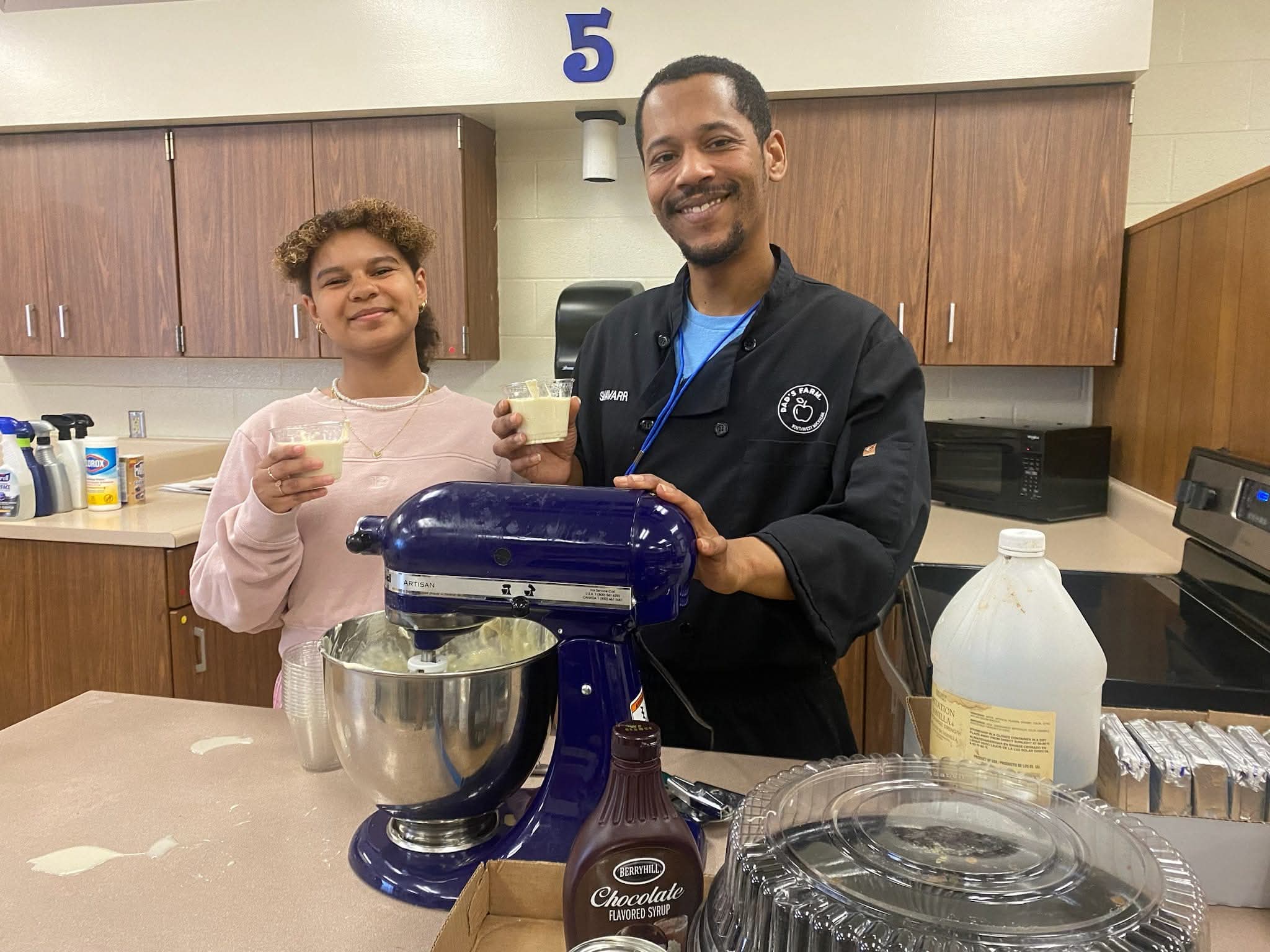 Two people smiling in a kitchen, holding cups, with a stand mixer and baking ingredients on the counter.