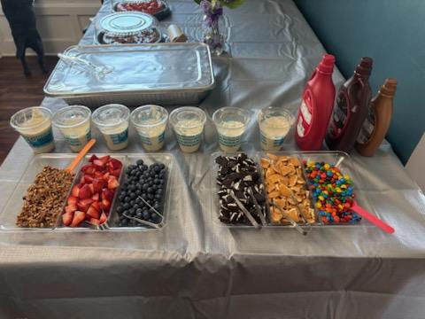 Table with dessert cups, toppings, and condiments, including strawberries, blueberries, chocolate, and colorful candies, on a white tablecloth.