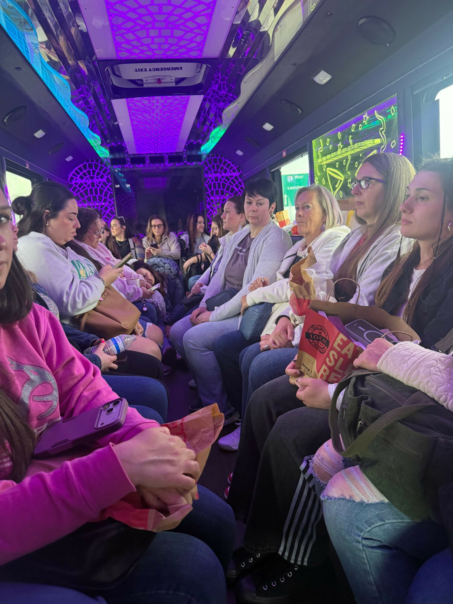 People seated in a row inside a bus or train, some holding snacks, with a digital display above showing colorful graphics.