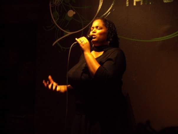 Woman singing into a microphone on stage with a dark background and decorative lighting.