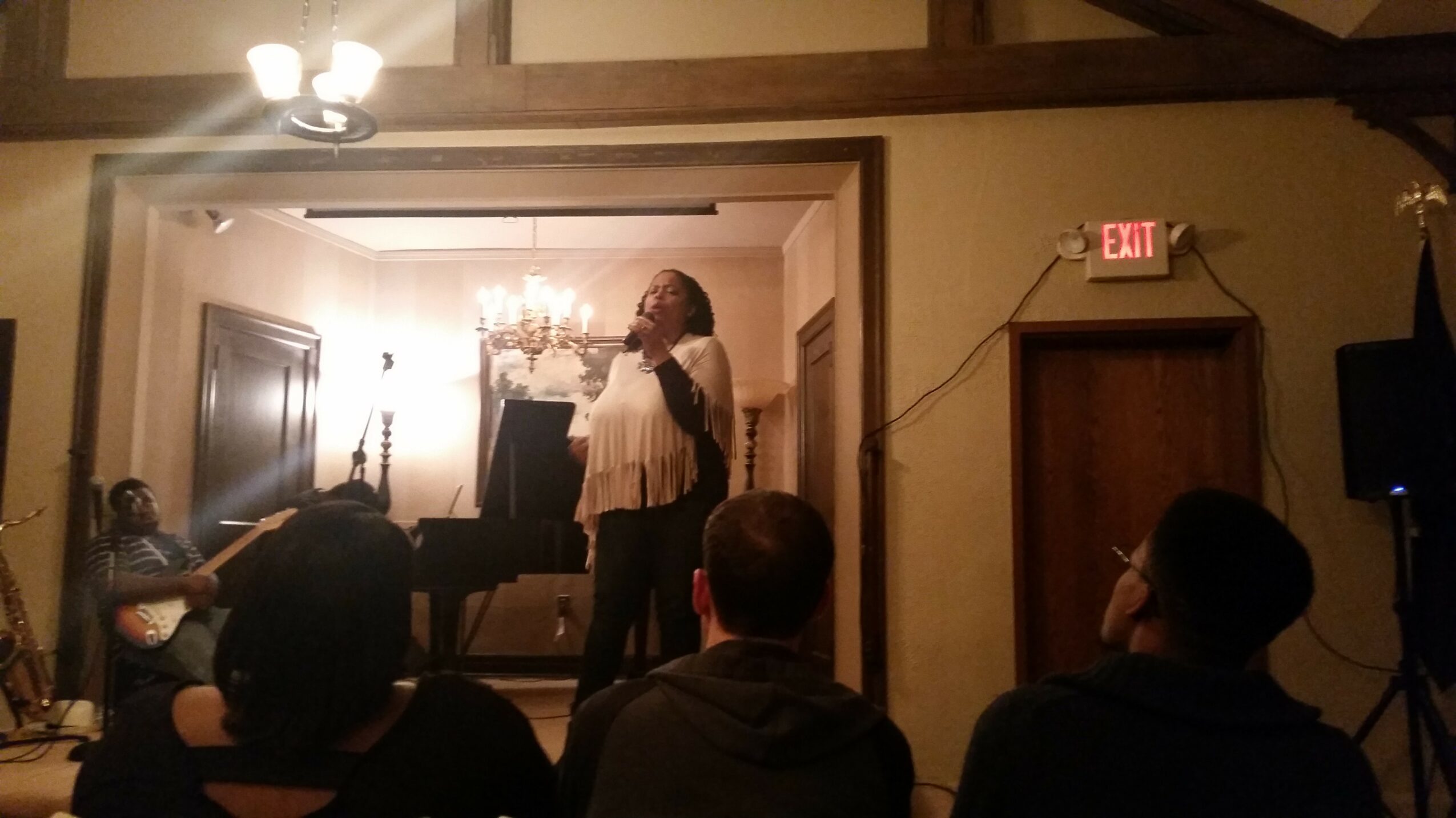 Person singing on stage with a microphone, audience seated watching, chandelier hanging, door with exit sign, warm lighting.