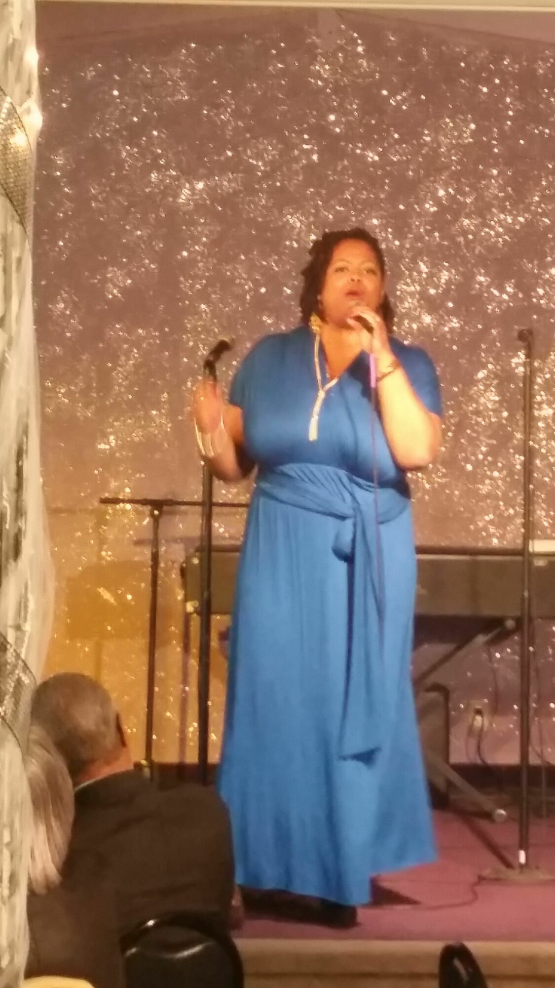 Woman in blue dress speaking into a microphone on stage with a starry backdrop.
