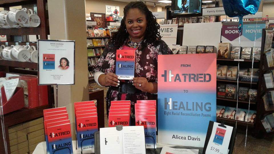 Woman standing behind a table with books and signs in a bookstore, smiling, holding a book. Books and signs are visible around her.