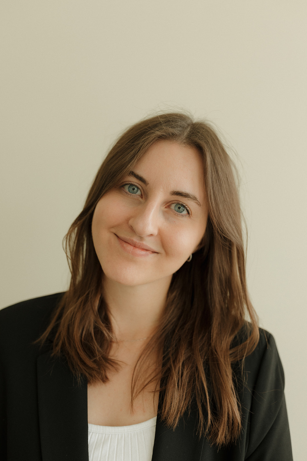 Young woman with long brown hair and blue eyes smiling, wearing a black blazer and white top, against a plain background.