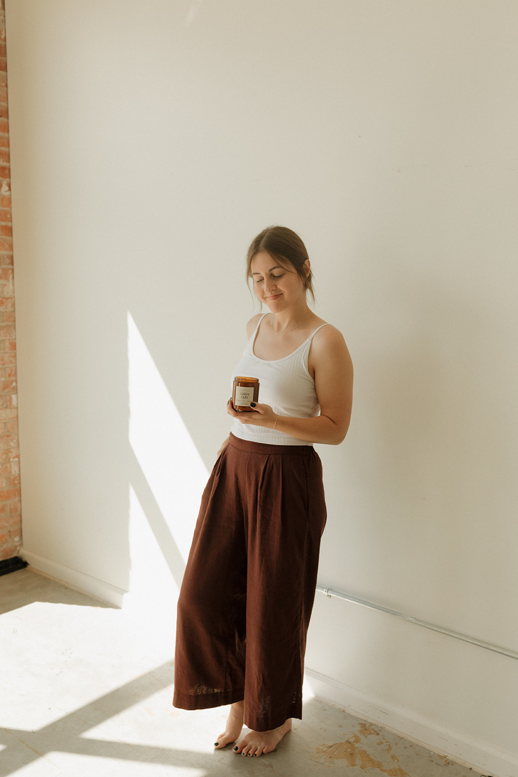 Young woman standing barefoot, reading a book, with sunlight casting shadows on a plain white wall.