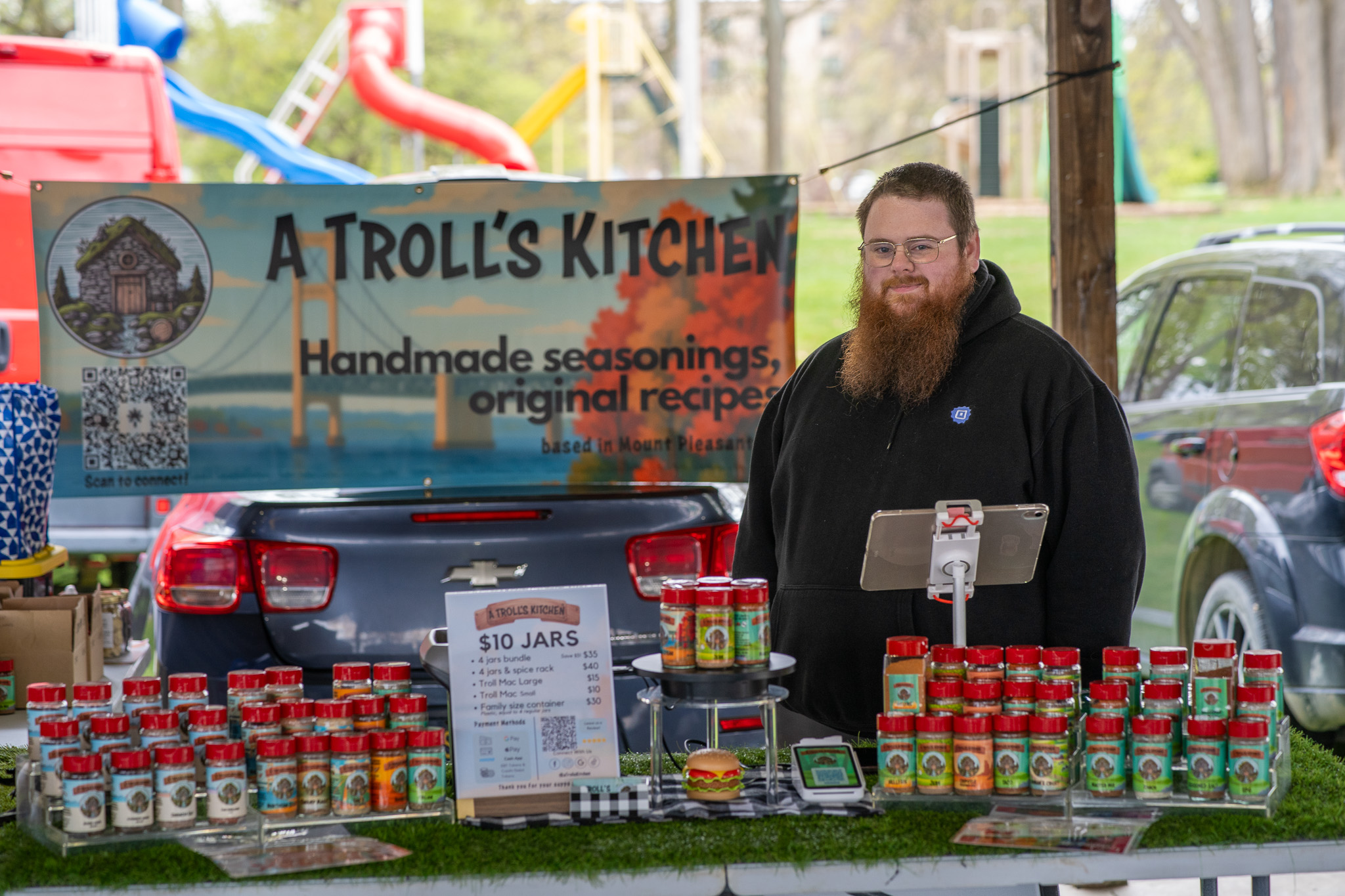 Man with a beard standing behind a food stall with jars and a sign that reads 'A Troll's Kitchen'.