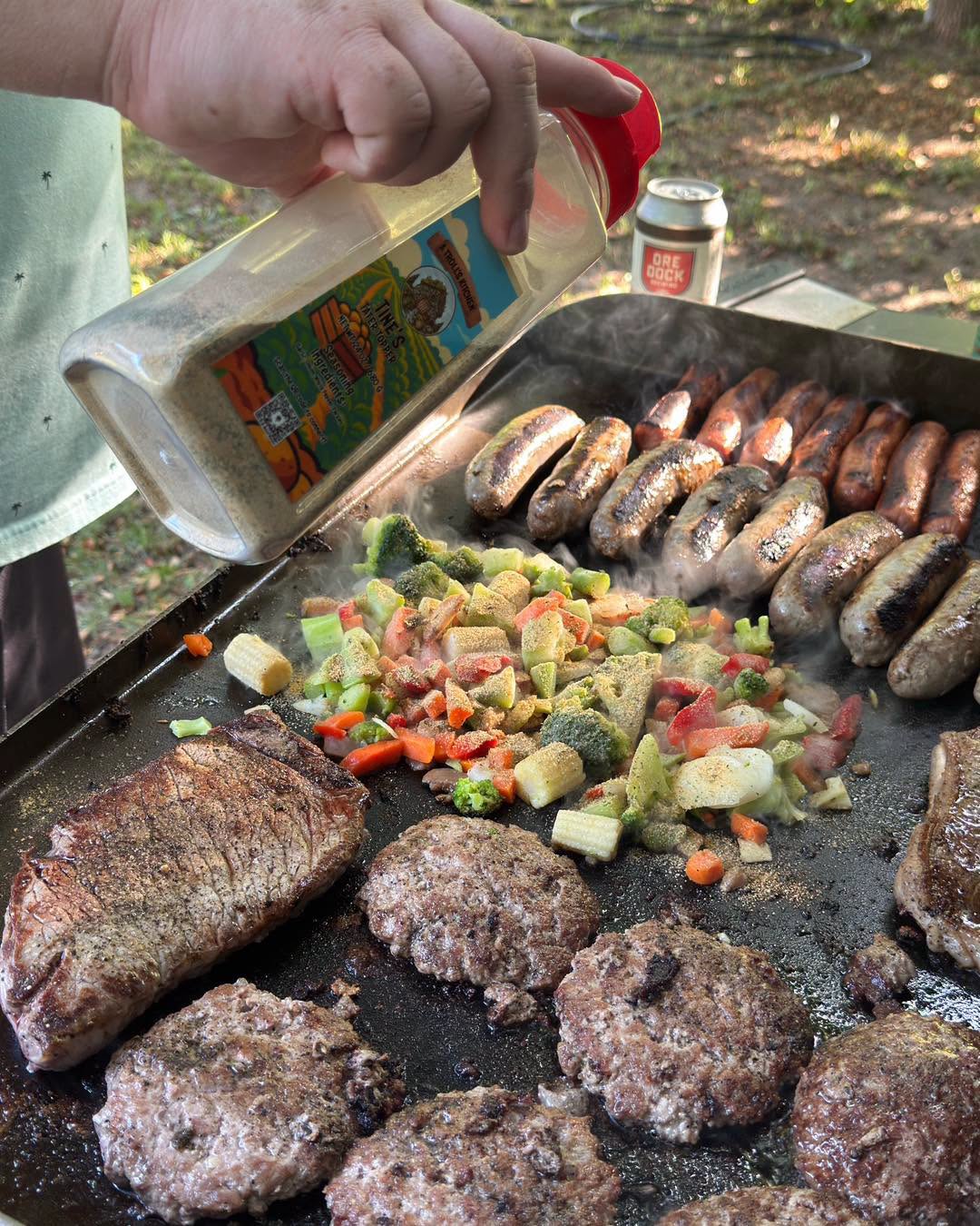 Person pouring seasoning on grilled sausages, vegetables, and burgers on outdoor barbecue grill.
