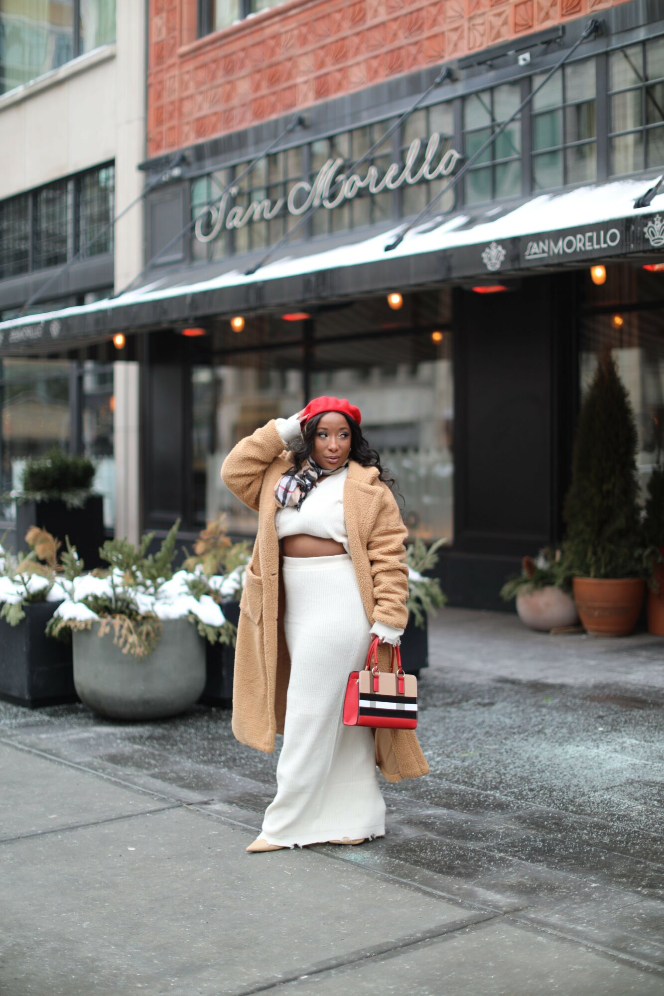 Woman in white dress and tan coat holding a red handbag standing on sidewalk outside a building with glass windows.