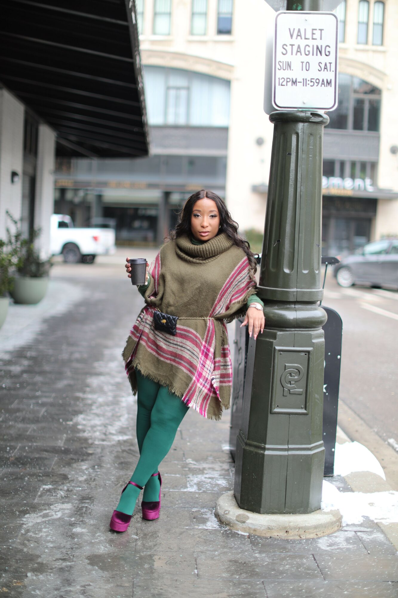Woman standing on sidewalk near a green parking meter, wearing a beige coat, plaid scarf, green pants, and pink shoes.